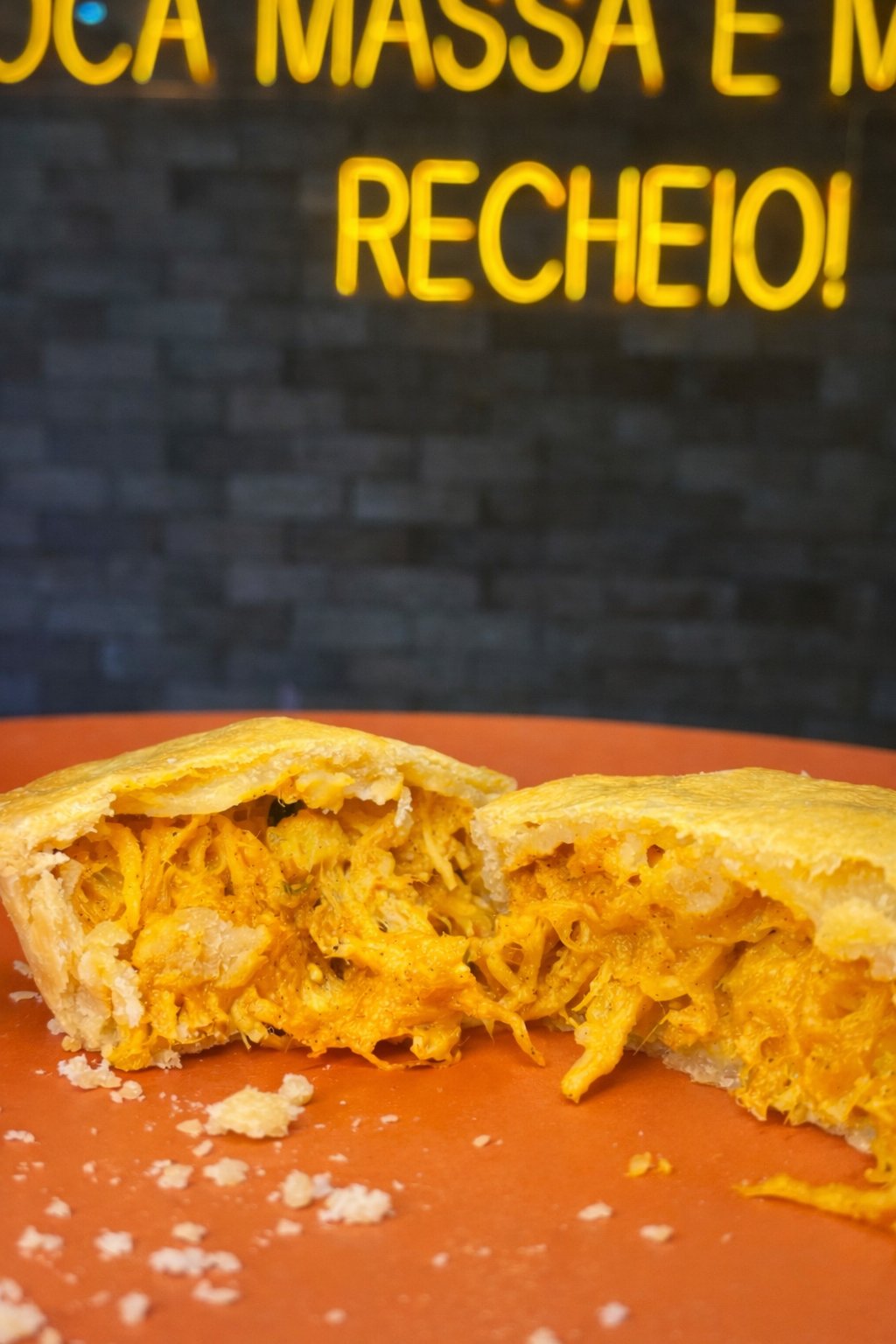 Empada de frango aberta ao meio com recheio generoso, diante do neon 'muito recheio' da Empada da Casa